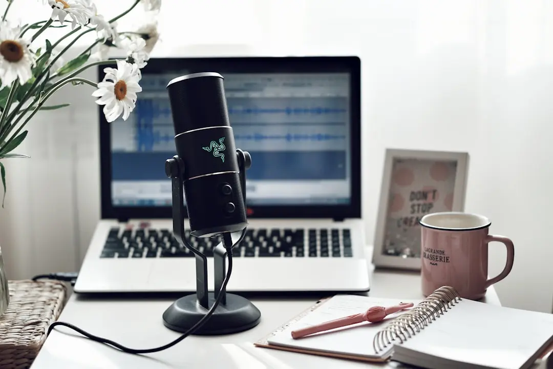 A microphone set up in front of a laptop with audio editing software on the screen, alongside a notepad, pen, and a mug on a home office desk for mastering blogging tasks.